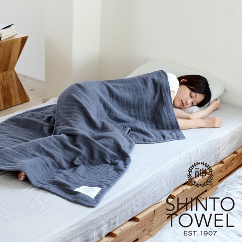 一位女性蓋著深藍色2.5層紗浴巾，浴巾質地柔軟，呈現自然的皺褶感，上有白色標籤，整體氛圍寧靜舒適。