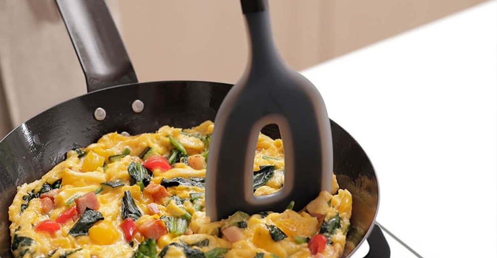 烹飪中的煎蛋捲，黑色鍋子裡有蛋、蔬菜和培根，其中一把黑色矽膠刮刀正在翻動食物。