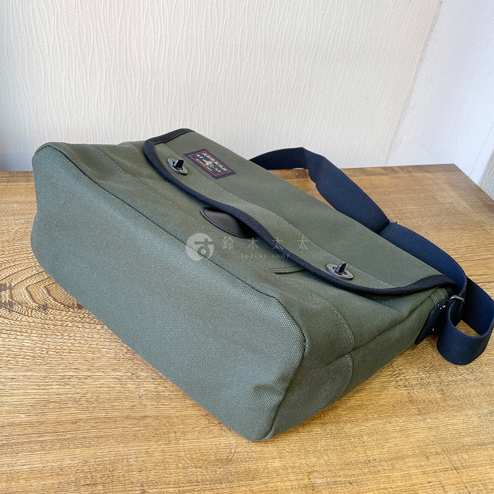 軍綠色純棉帆布雙扣郵差包，帶有黑色邊緣裝飾和黑色肩帶。包蓋上印有紅色標籤，並配有兩個金屬扣。