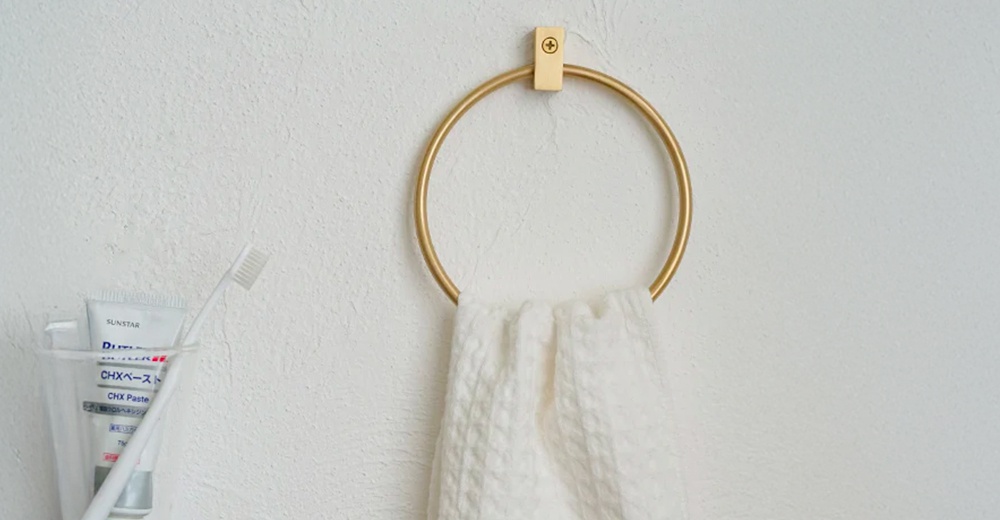 金色黃銅圓形毛巾環，掛在白色紋理牆面上，環上掛著一條白色紋理毛巾。