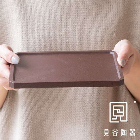 一雙手捧著一個長方形的深棕色烤盤，烤盤邊緣有斜角設計，表面光滑無紋理。