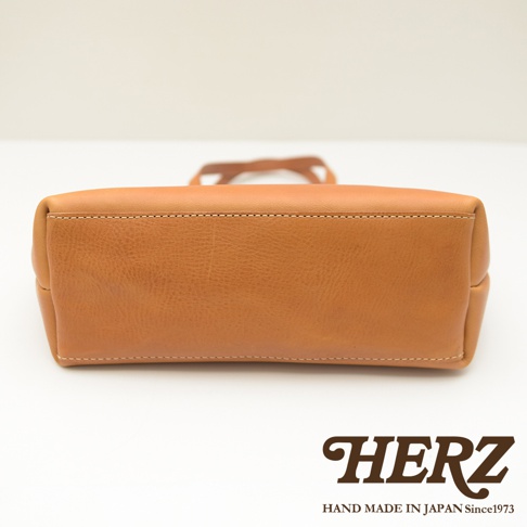 職人手作皮革托特包，呈現溫潤的淺棕色，材質為真皮，包體底部邊緣有細緻的白色縫線，整體設計簡約。