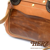 職人手作皮革郵差包，採用天然皮革製成，呈現棕色漸層紋理，以縫線和金屬釦點綴，設計簡約實用。