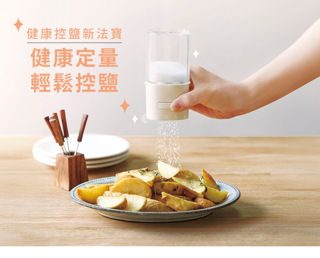 一隻手正拿著白色定量灑鹽罐，罐身為米白色，上方為透明玻璃材質，裡面裝有白色細鹽，灑出細鹽於盤中的烤馬鈴薯上。