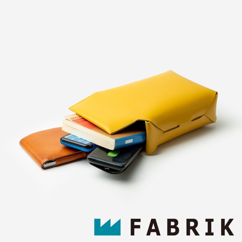 FABRIK 品牌標誌旁是黃色皮革斜背包，裡面裝著手機和書本，旁邊還有一個橘色皮夾