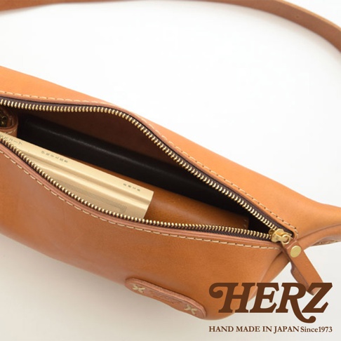 職人手作皮革輕便斜背包，採用棕色皮革製成，拉鍊打開，內部可見一本書和一個長形物品。