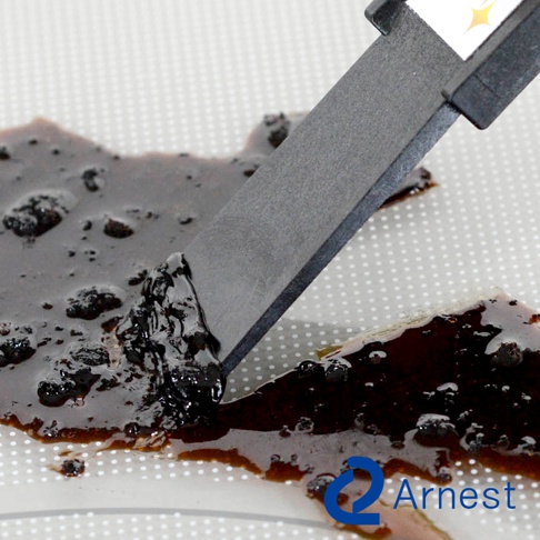 灰黑色刮刀正在刮除深棕色黏稠物，刮刀邊緣呈現磨砂質感，背景為淺灰色帶有白色圓點的平面。