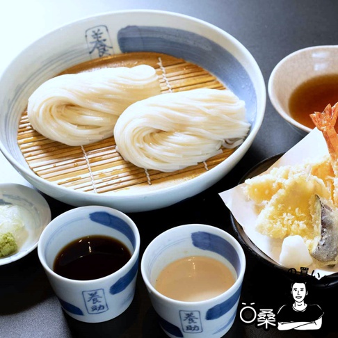 兩份白色的稻庭烏龍麵擺放在竹篩上，旁邊配有炸物（包含炸蝦和蔬菜）、醬汁和芥末。