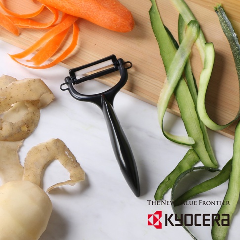 黑色陶瓷削皮刀，刀頭為白色陶瓷，配有符合人體工學的黑色手柄，旁邊有已削皮的馬鈴薯、胡蘿蔔和黃瓜。