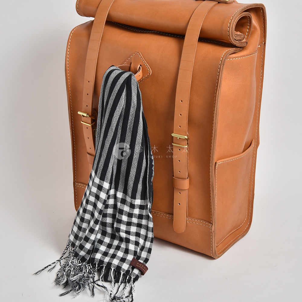 一款棕色皮革後背包，配有可調節肩帶和黃銅扣環，背包一側掛有一條黑白格紋圍巾，圍巾帶有流蘇。