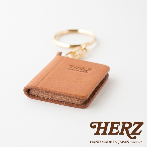 職人手作皮革書本造型鑰匙圈，呈現淺棕色皮革，搭配金色鑰匙圈，書本封面壓印有「HERZ」品牌標誌。