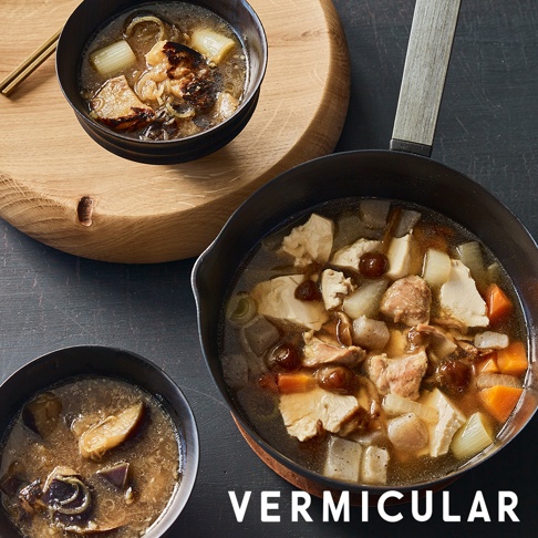 畫面中央為一只黑色 VERMICULAR 琺瑯鑄鐵雪平鍋，鍋內盛裝著含有雞肉、豆腐、紅蘿蔔及蒟蒻的豐盛燉煮料理，兩側各放置一個黑色小碗，裝有同樣烹調風格的湯品，上方木質砧板擺放著一雙筷子，整體呈現出溫暖居家的料理情境。