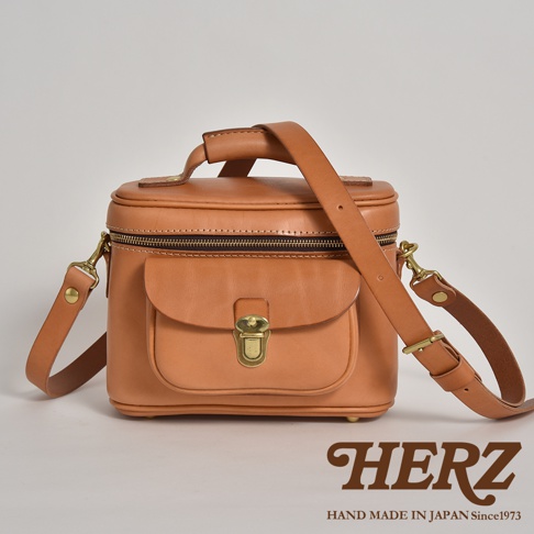 職人手作的淺棕色皮革相機包，方形立體設計，頂部有提把，正面有翻蓋口袋，以黃銅扣環固定，側邊有可拆卸的長背帶，整體風格簡約復古。