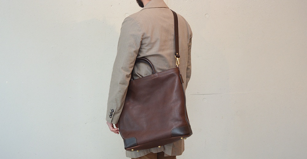 一位男士背著一個深棕色皮革托特包，包包的底部有金屬裝飾。