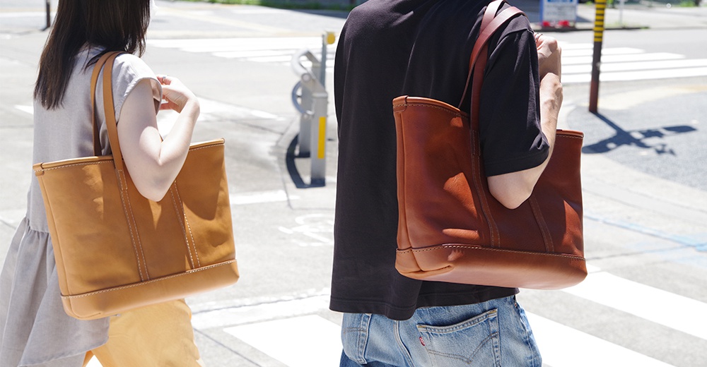 兩位人士各持一個皮革托特包，左邊為淺棕色，右邊為深棕色。包款設計簡約，採用皮革材質，配有提把，展現經典風格。