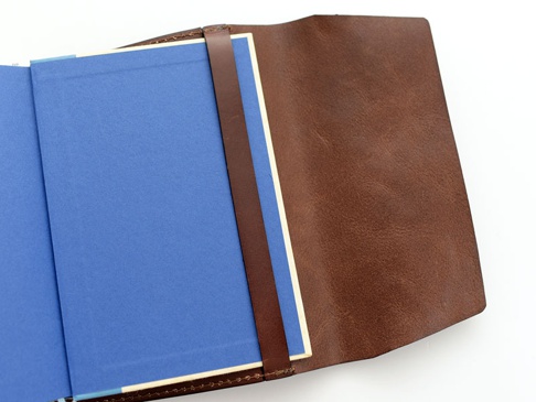 棕色皮革翻蓋記事本書套，內頁為藍色，書套以一條同色皮革帶固定，整體呈現簡約質感。