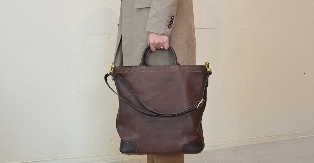一名穿著米色外套的人手提一個深棕色皮革托特包，包上有可拆卸肩帶，包的底部有黑色皮革加固。