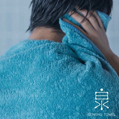 特寫鏡頭顯示一件藍綠色毛巾，質地蓬鬆柔軟，一位人士正用毛巾擦拭臉部。毛巾邊緣有藍色滾邊。