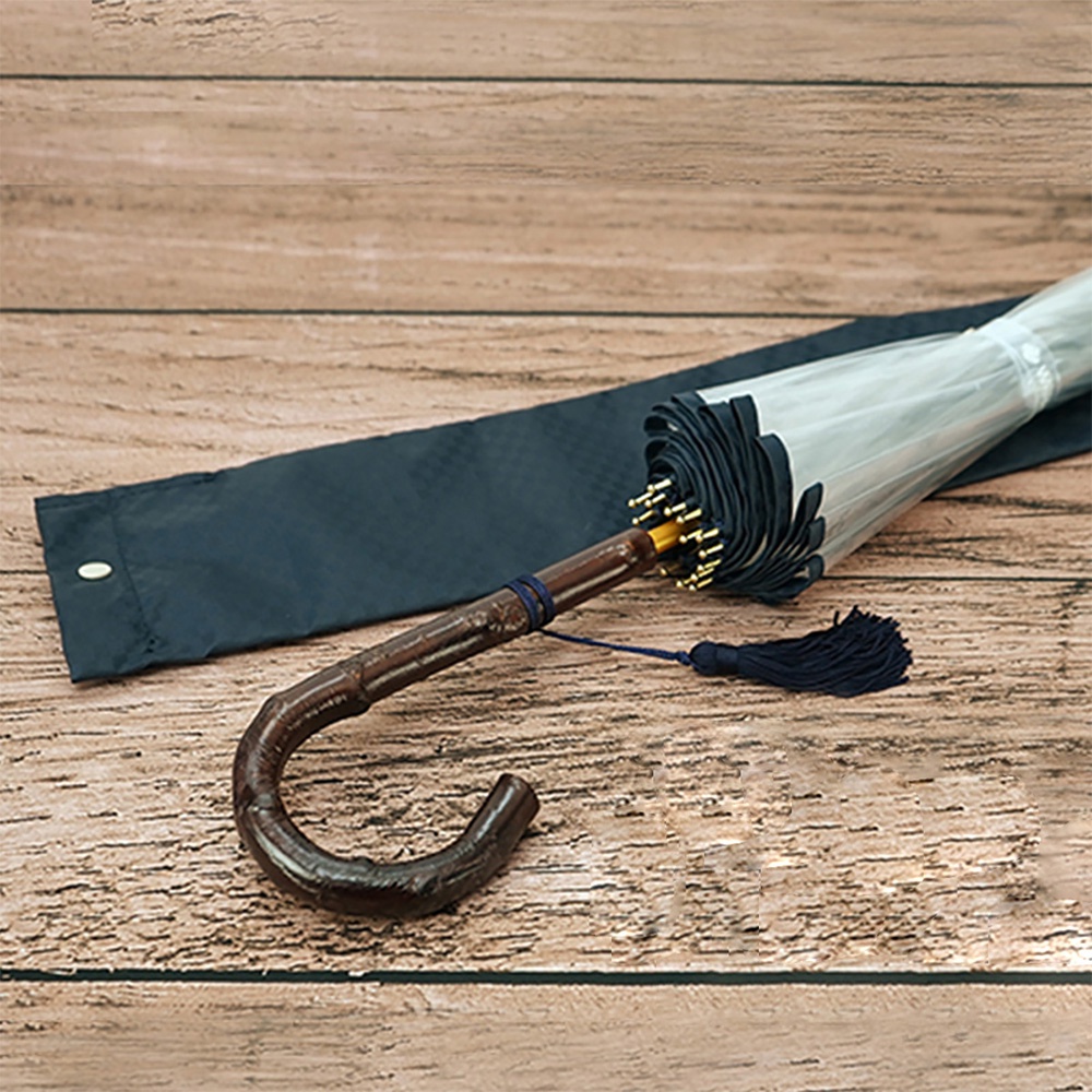 一把半開的透明傘，傘骨為深棕色彎曲木質握把，傘柄束有深藍色流蘇，配有深藍色傘套，置於木紋背景上。