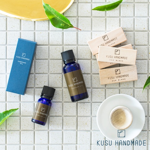 藍色玻璃瓶裝的樟楠木精油，瓶身標有「KUSU HANDMADE CAMPHOR OIL」字樣，旁邊擺放著藍色紙盒、堆疊的淺木色樟楠木塊，以及一個白色小碟。