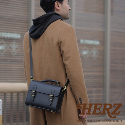 黑色皮革迷你兩用包，帶有兩個黃銅搭扣和頂部提把，以斜肩包形式展示，由穿著駱駝色大衣的男性攜帶。