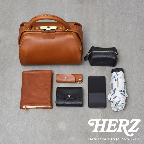 職人手作皮革Organ兩用口金手提包 G-81，以棕色皮革製成，包身硬挺，搭配金色金屬鎖扣與提把。旁邊擺放著黑色皮革零錢包、棕色皮革筆記本、黑色皮革名片夾、手機和摺疊傘。