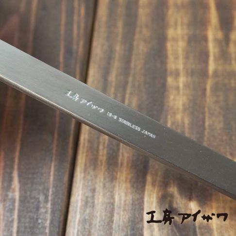 銀色不鏽鋼萬用瀝水湯勺的特寫，勺柄上刻有日文「工房アイザワ」和「18-8 STAINLESS JAPAN」字樣，背景為木紋桌面。