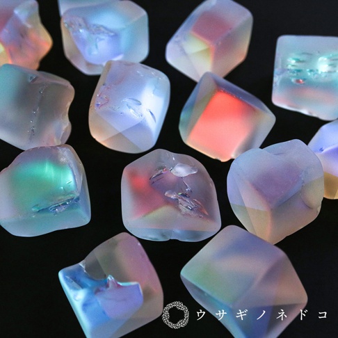 一群不規則形狀的透明稜鏡石，表面光滑，內部折射出粉紅、藍、綠、黃等色彩光芒，部分石塊內嵌有細小的裝飾物。