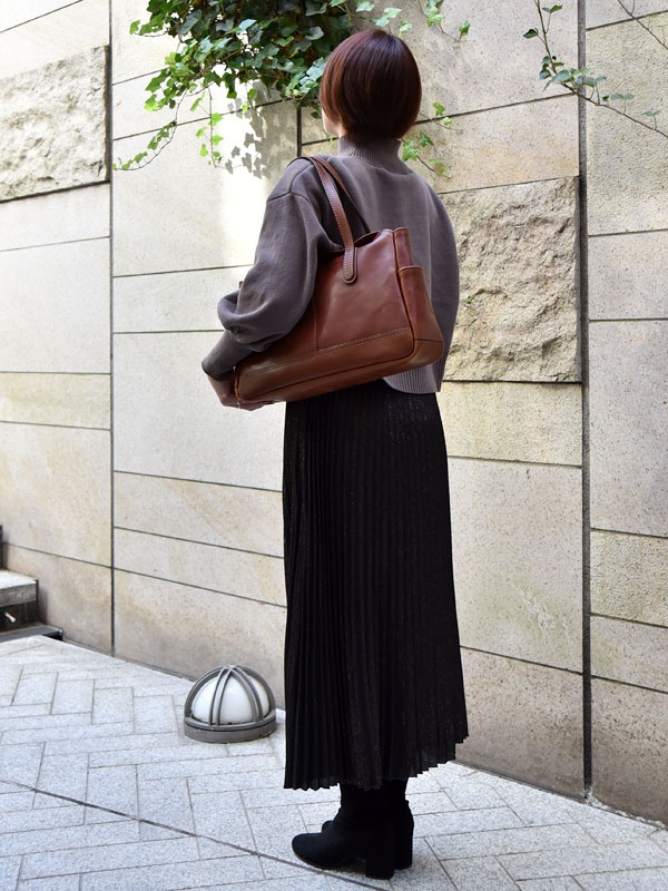 一位女性手提棕色皮革製的橫式托特包，包包帶有縫線細節，側面有兩個小口袋。