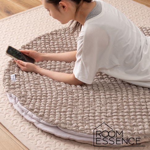 一位女性趴在一個淺褐色、帶有立體紋路的圓形涼感墊上，雙手持手機。涼感墊下方露出白色內襯。