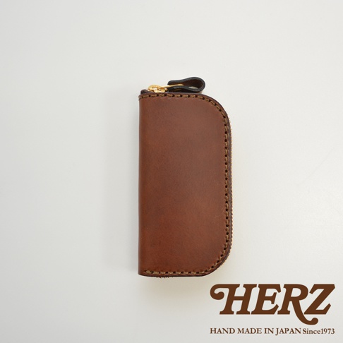 一個棕色皮革製成的橢圓形鑰匙包，頂部有金色拉鍊和黑色皮革拉環，側面有縫線細節。底部有 HERZ 品牌標誌。