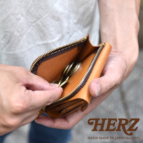 一雙手正拿著一個日本HERZ品牌職人手作的淺棕色皮革短夾，拉鍊開口處清晰可見內部的零錢，整體呈現出質感優雅的皮革紋理與精緻工藝細節。