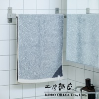 一條淺灰色棉麻混紡毛巾，底部有米白色滾邊，右下角有深藍色幾何圖案。毛巾掛在銀色金屬毛巾架上，背景為白色磁磚牆。