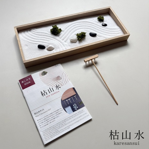 這是一組日本枯山水模型套件，包含一個長方形木製托盤、用於裝飾的白色細沙、數顆黑色與白色石頭、幾叢模擬青苔裝飾，以及一把木製耙子。前方擺放一本印有日文與英文介紹的說明書，封面顯示著該套件的組件排列範例，整體呈現出簡潔、禪意的日式庭園風格。