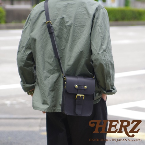 一位背著日本HERZ職人手作黑色硬皮革單肩包的男子，包款採用經典皮帶扣環設計並搭配黃銅五金配件，呈現簡約俐落的背影視覺，下方標註品牌名稱與產地資訊。