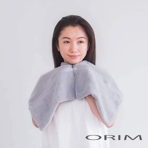 一位女士身穿一件淺灰色、柔軟觸感的吸水毛巾，設計成披肩形狀，可固定於頸部，方便吹乾頭髮。
