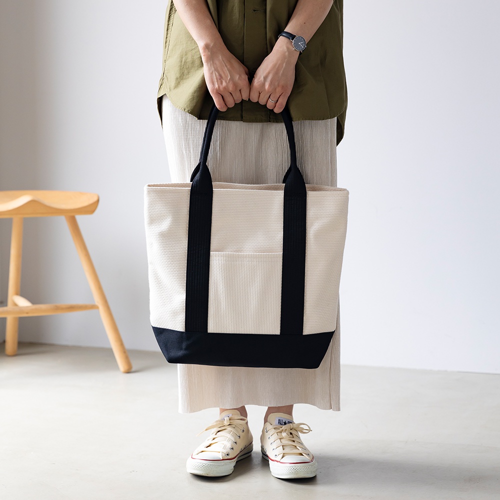 米白色三河木綿刺子織托特包，配有黑色織帶提把及底部，正面有一個黑色貼袋。