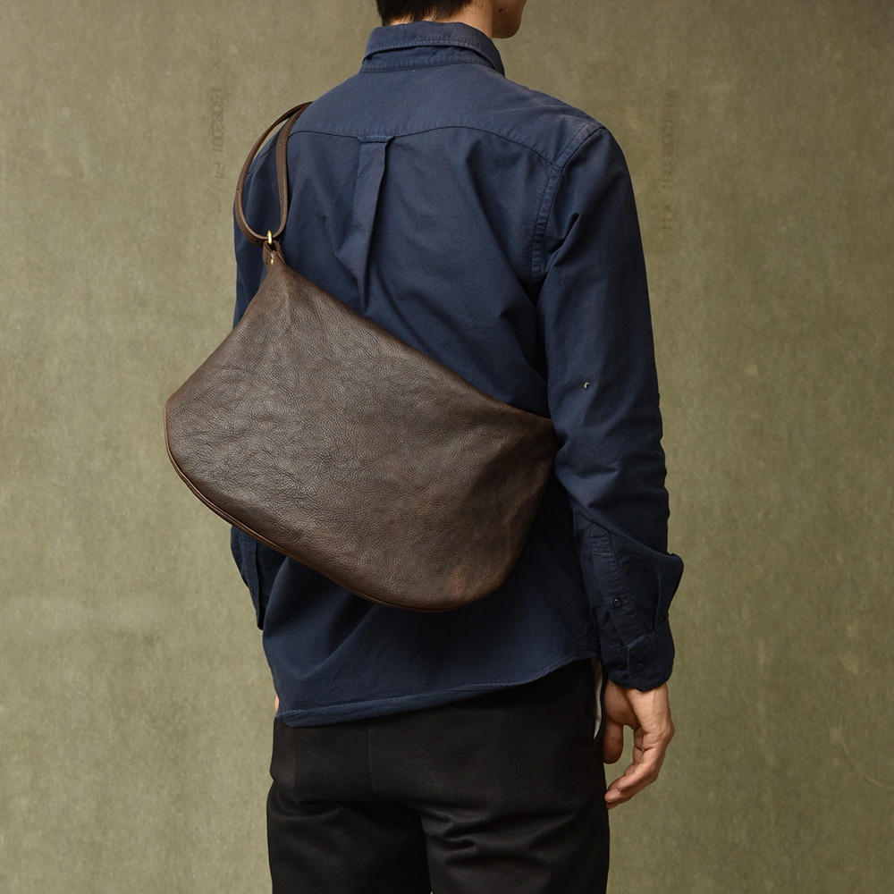 深棕色職人手作皮革Organ單肩半月包，以自然紋理皮革製成，半月形設計，斜跨於身著海軍藍襯衫的模特身上，展現簡約質感。