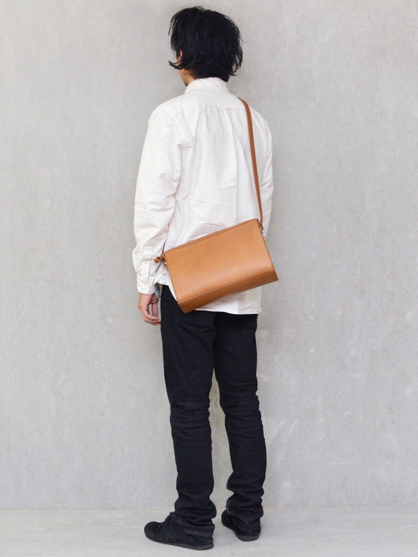 皮革製的棕色拉鍊單肩包，以簡約設計斜挎於身穿白色襯衫和黑色長褲的模特肩上。