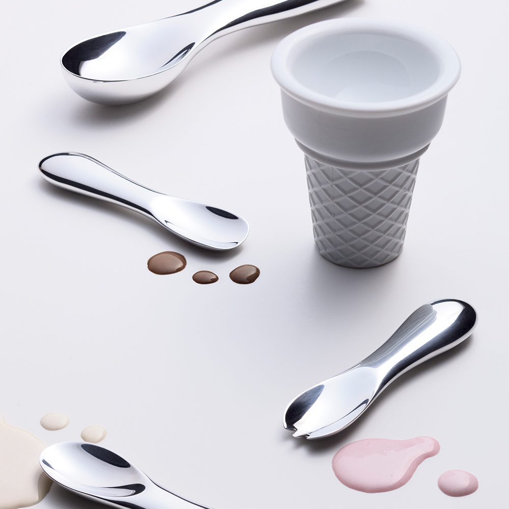 四款金屬冰淇淋勺，其中兩款為純勺形，一款為帶有叉齒的勺子，另一款為較大的勺子。旁邊有一個白色陶瓷製成的冰淇淋甜筒形狀的容器，表面飾有菱格紋。背景中有幾灘不同顏色的液體，像是巧克力醬、香草醬和草莓醬。