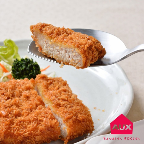 銀色金屬切切分食匙，夾著一片炸豬排，豬排外層金黃酥脆，內層肉質粉嫩。盤中還有其他炸豬排塊，搭配生菜、紅蘿蔔絲及花椰菜。