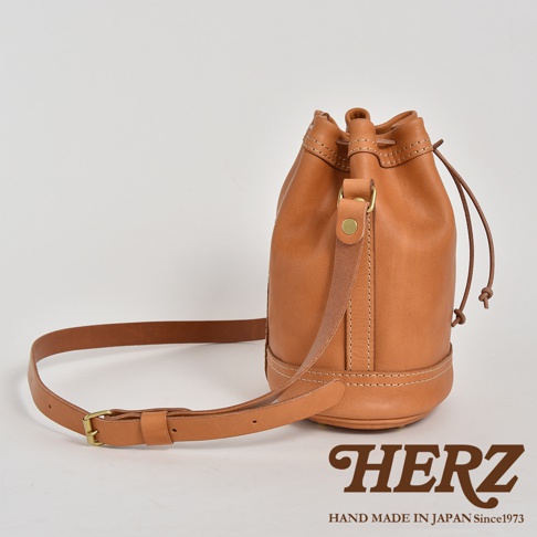 職人手作的棕色皮革束口單肩包，圓筒形包身，附有可調節長度的皮革背帶和束口抽繩，包身下方有縫線裝飾。