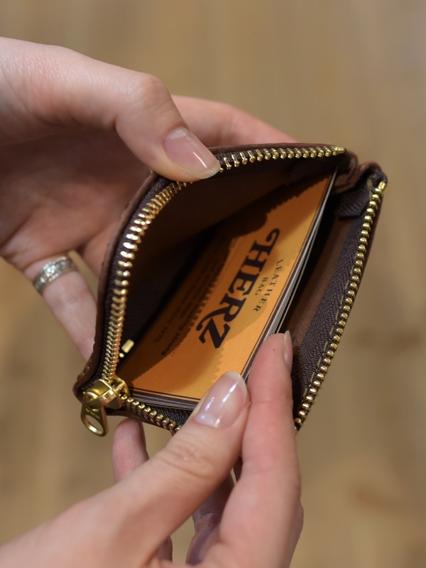 深棕色軟皮革L型拉鍊卡片夾，內有數張印有「HERZ」字樣的黃色名片，由雙手正在開啟。