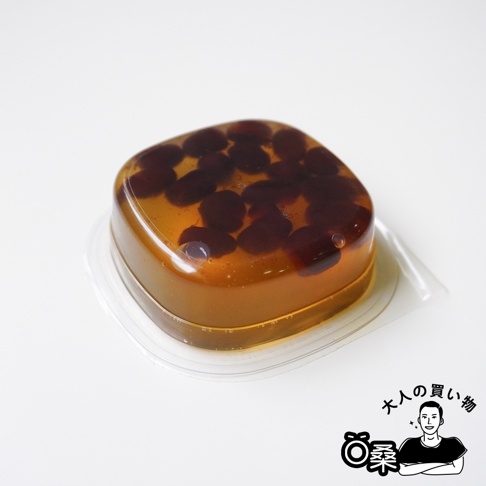 放置在透明塑料盒內的方形紅豆蕨餅，呈半透明琥珀色，外觀光滑且充滿彈性，清晰可見內部包裹的深色紅豆顆粒，右下角帶有大人的買物標誌。