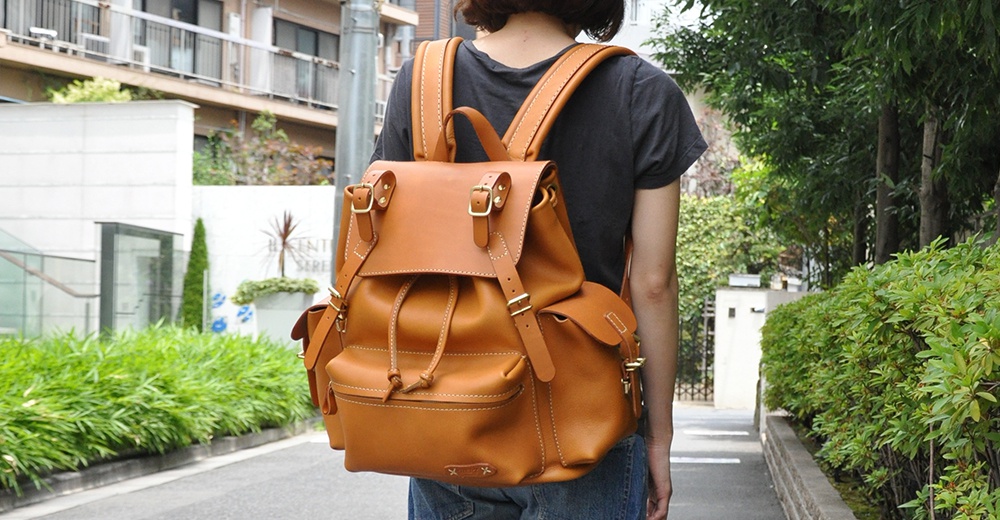 一個棕褐色的皮革旅行後背包，有著復古風格的設計，包含抽繩開口、翻蓋式閉合、兩個側邊口袋和可調節的肩帶。