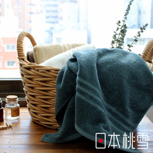 藤編籃中疊放著今治飯店浴巾，包括深綠色、白色和米色。其中一條深綠色浴巾搭在籃子邊緣，帶有織紋裝飾。