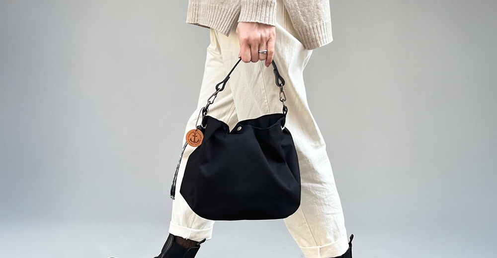 一位女性手提著神戶clueto的黑色BALON mini防潑水手提包，包身圓潤簡約，並掛有一個帶有船錨圖案的圓形木質吊牌，展現出俐落時尚的穿搭風格。