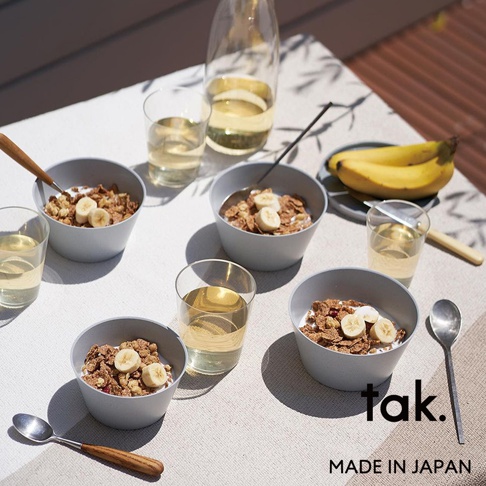 四個淺灰色餐碗，內有麥片、牛奶和香蕉切片，搭配玻璃杯裝的飲品及木質湯匙，置於白色桌布上。