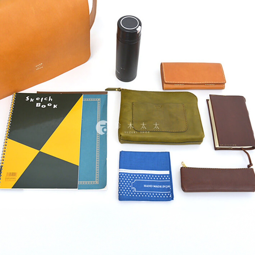 圖片內有「Sketch Book」、「SUZUKI SHOP」、「HAND MADE IN JAP」等文字，整體展示了多款手工皮革製品，包含一個棕色托特包、一個黑色保溫瓶、一個橘色皮夾、一個綠色化妝包、一本筆記本、一個藍色手帕和一個棕色長夾。