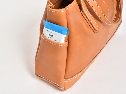 淺棕色皮革橫式肩背托特包，側邊外袋置放一包藍色包裝的濕紙巾。包包以白色縫線裝飾，呈現職人手作質感。